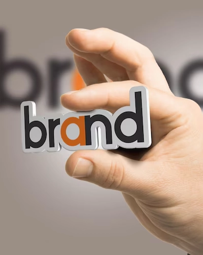 A hand holding a 'brand' word which is in black & orange colour,'a' letter alone coloured in orange