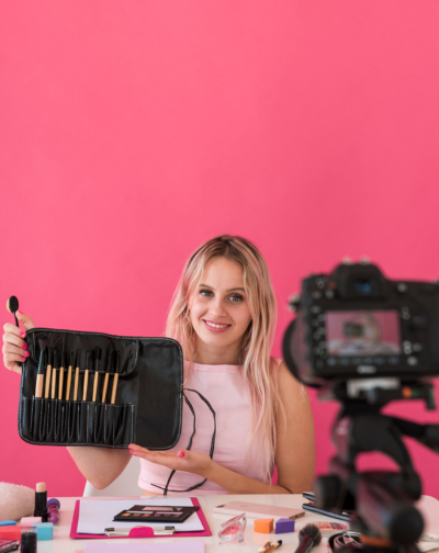 A woman holding a digital camera and makeup brushes, preparing to start her video business.